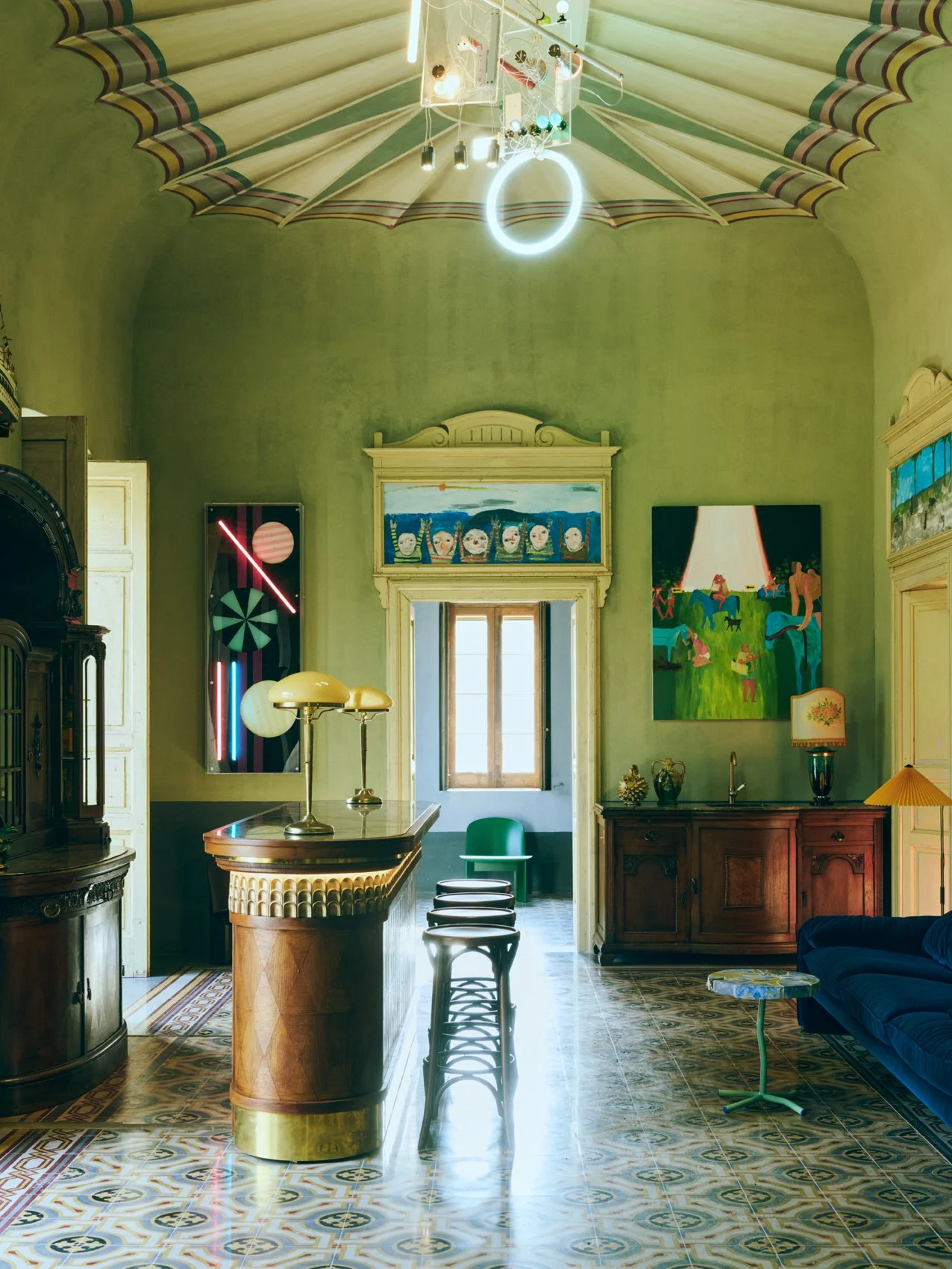 Interior of a vintage room with high ceilings, decorated with modern art and eclectic furniture, including a yellow bar counter, green chairs, a blue sofa, and colorful lighting fixtures, with brightly patterned tiled floors.