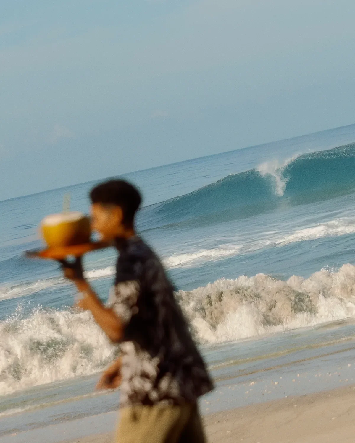 Person walking on the beach holding a coconut drink with a straw, with ocean waves and a surf beach in the background.