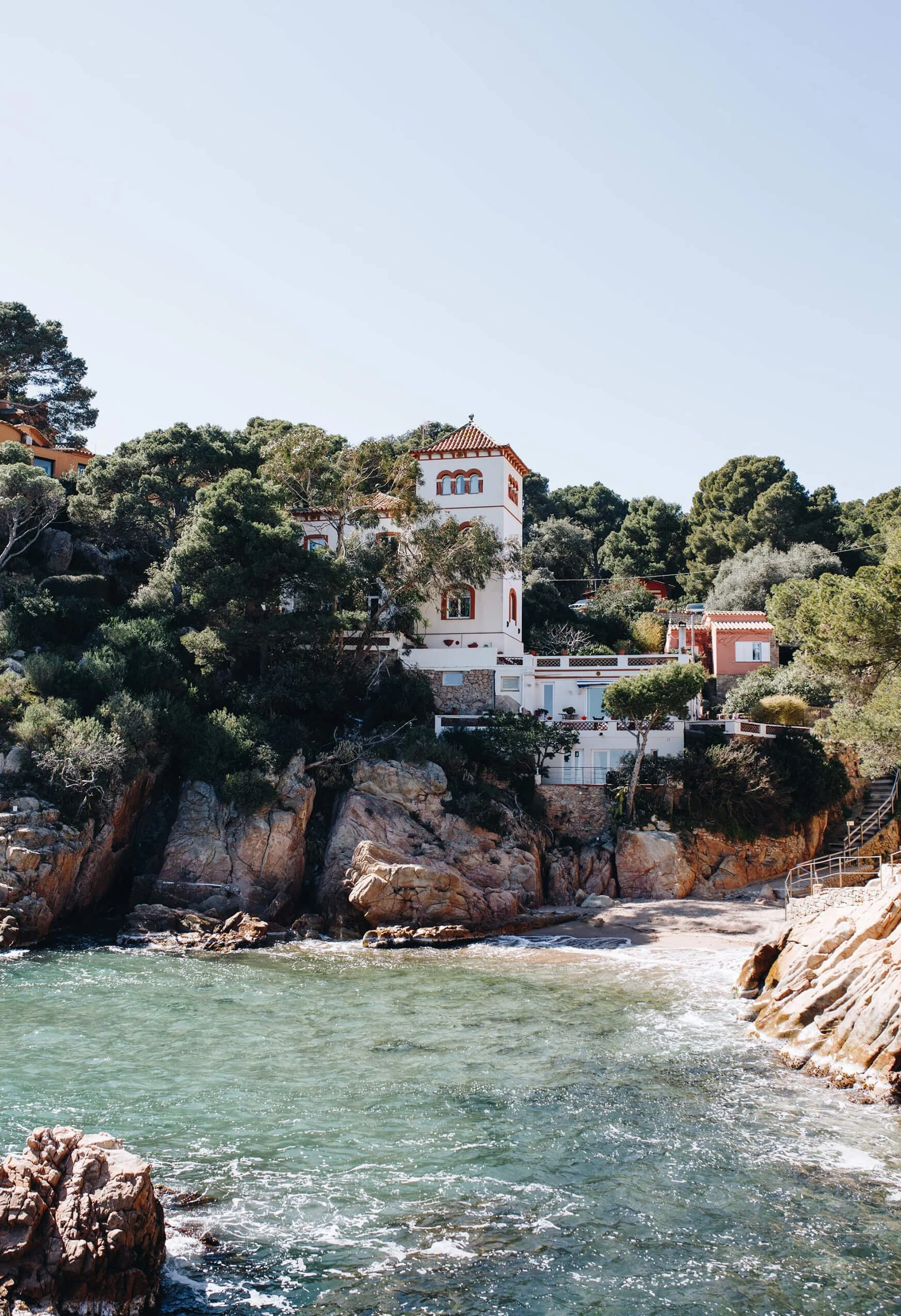 A coastal scene with a rocky beach in the foreground, calm water, and a hillside with white and red-tiled houses surrounded by trees.