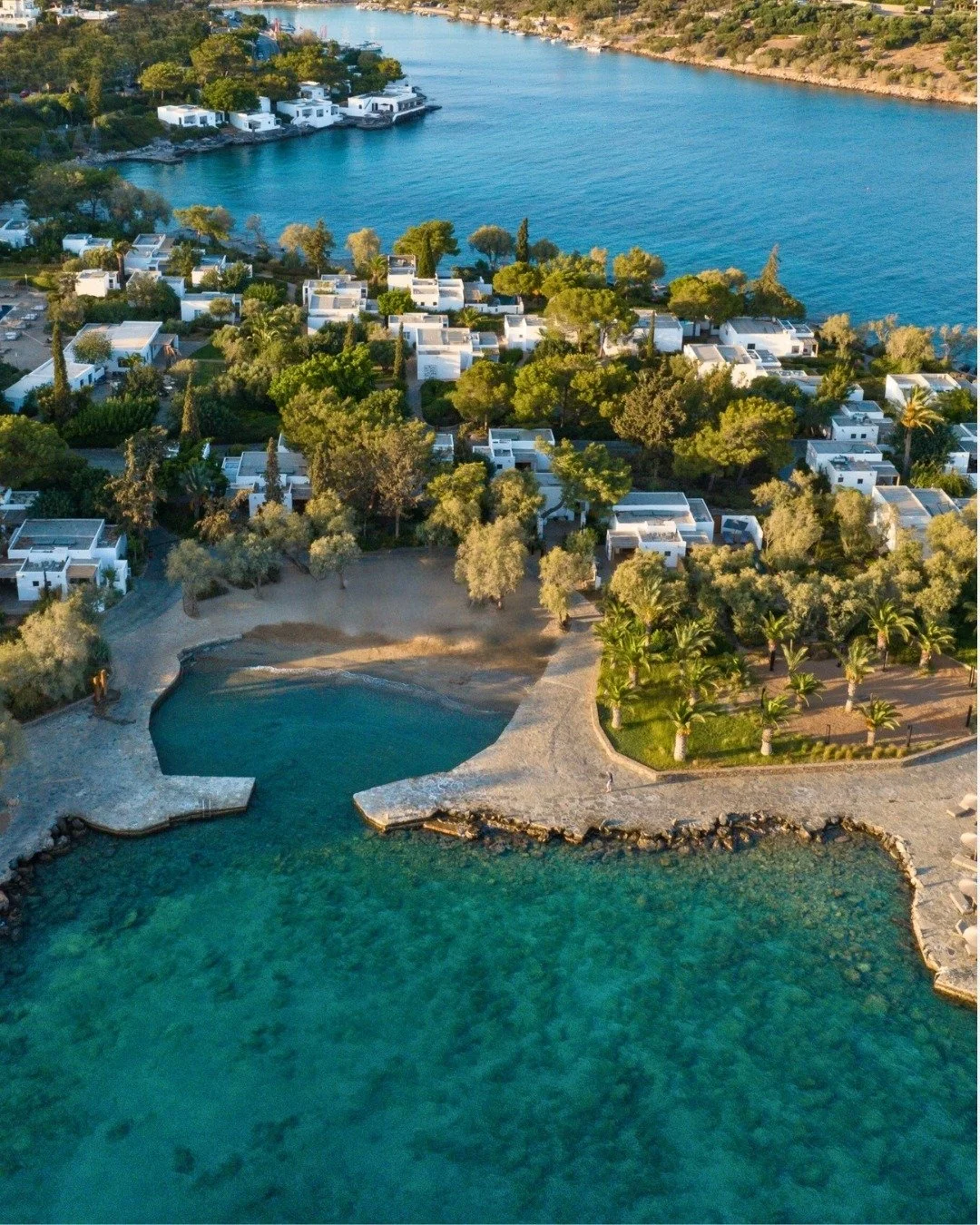 Aerial view of a coastal resort with white buildings, palm trees, a small beach, a bay with turquoise water, and boats docked along the shoreline.