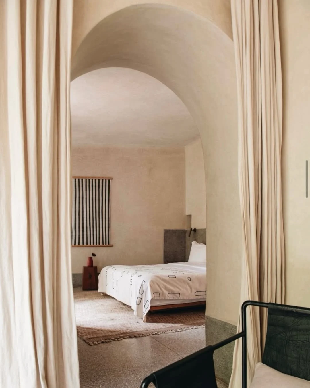 View of a bedroom through a curtain archway, showing a bed with white sheets, a nightstand with a red lamp, a window with a striped curtain, and a textured area rug.