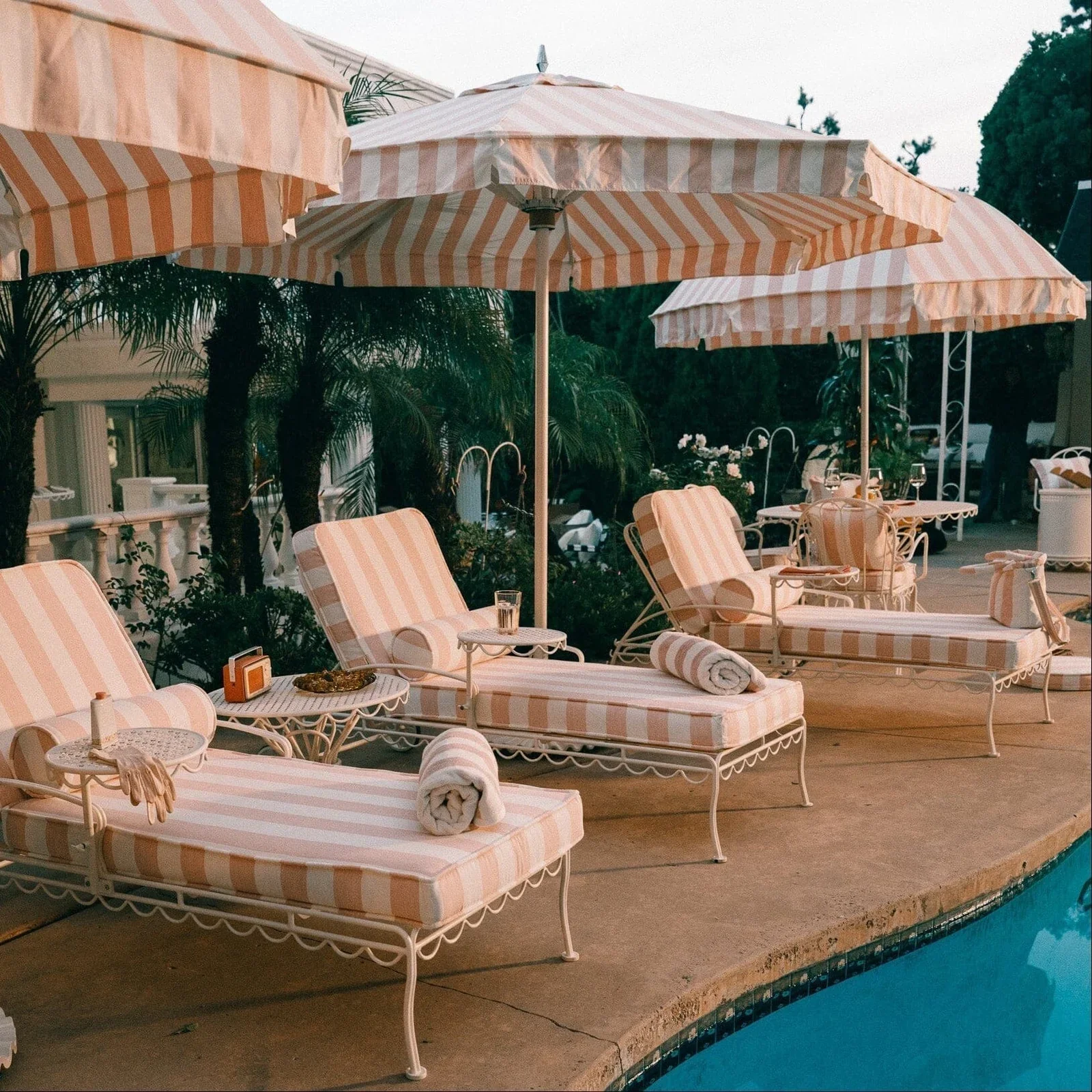 Poolside scene with striped pink and white lounge chairs, umbrellas, and tropical plants in the background.