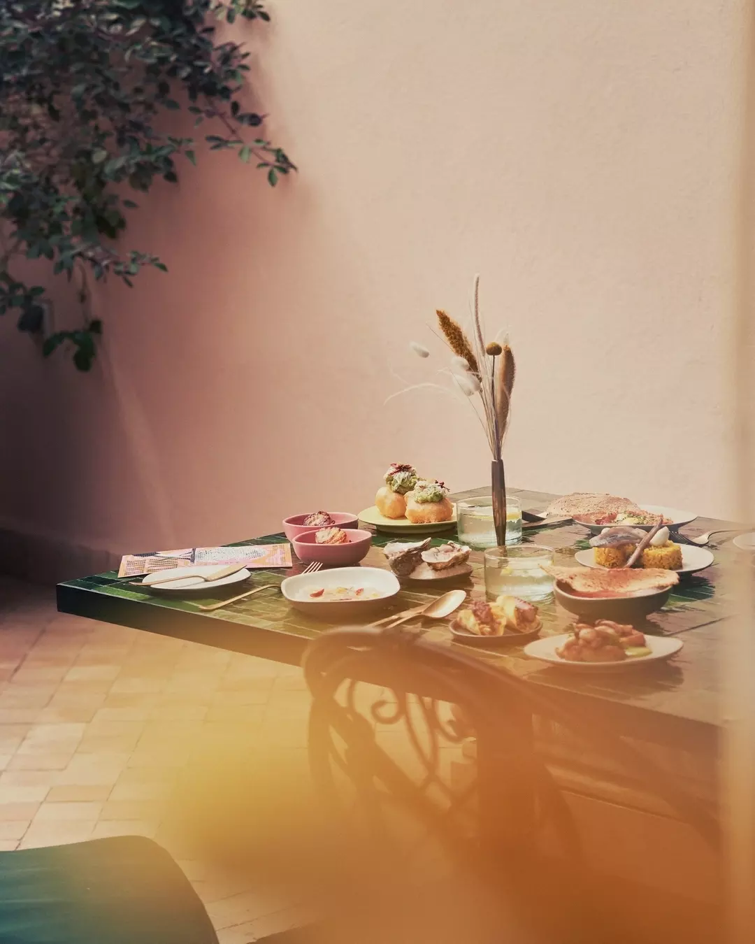 A dining table with various plates of food, a vase with dried flowers, and a nearby plant in a corner.