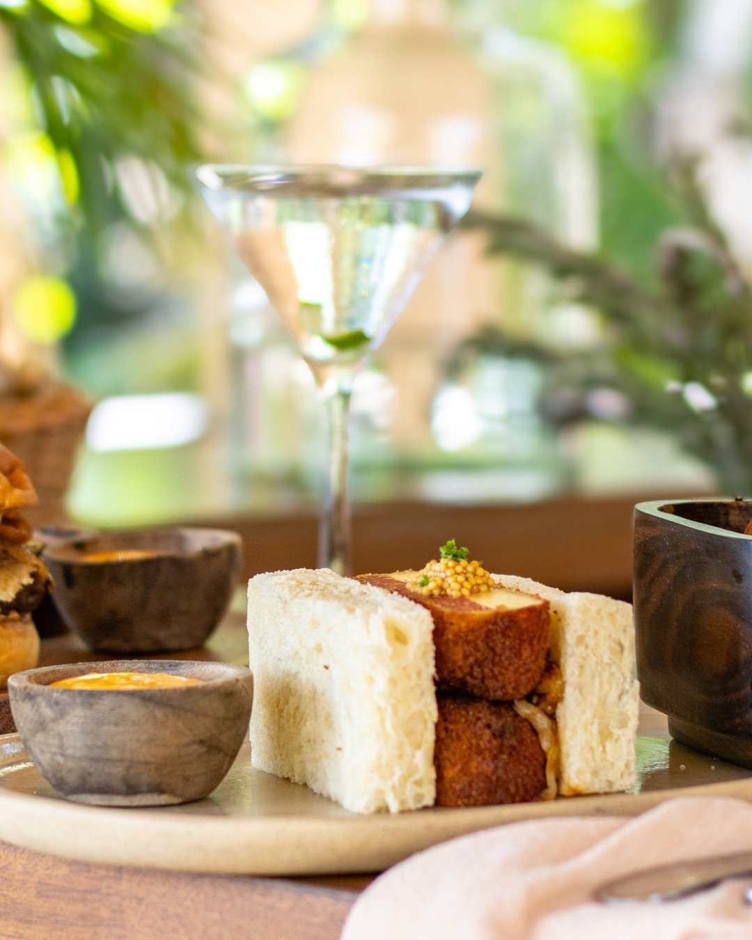 A close-up of a bread and cake platter with a glass of cocktail in the background, set on a wooden table in a bright, green outdoor setting.
