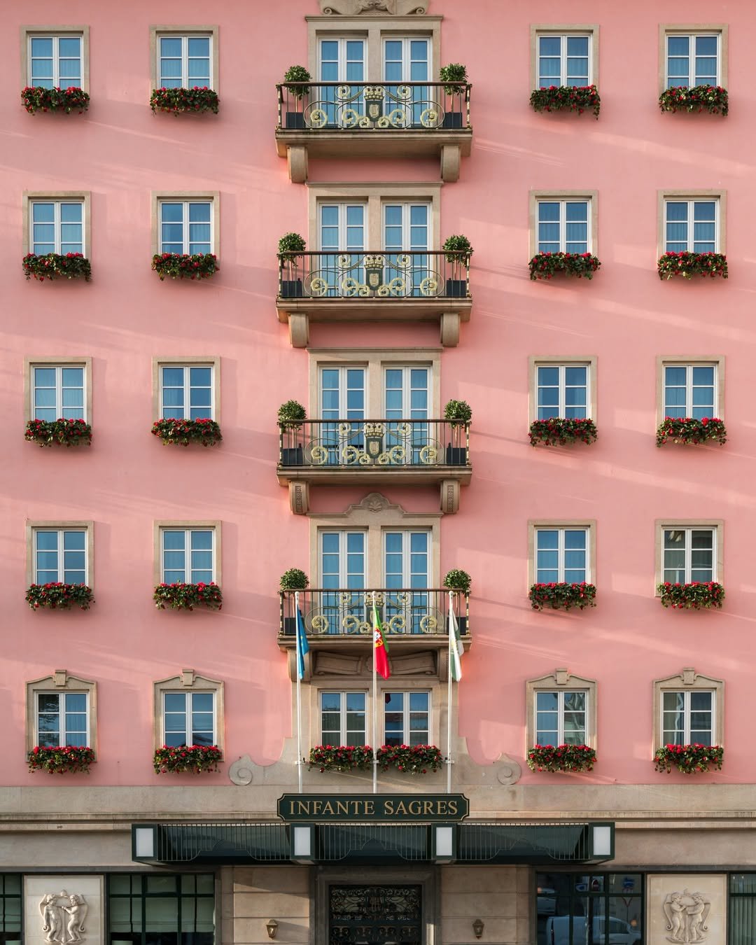 Pink building with multiple windows, each adorned with flower boxes filled with red flowers. Three balconies with decorative railings and small potted trees. Flags on poles in front of the building, and a sign reading "Infante Sagres" above the entrance.