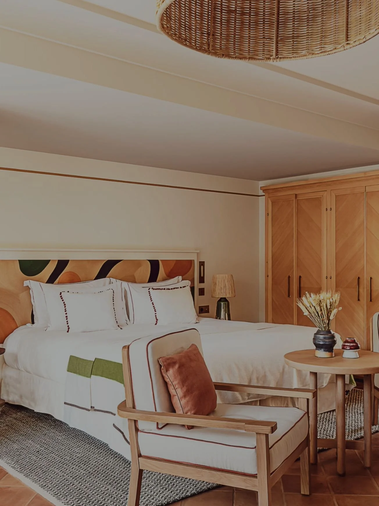 A cozy hotel bedroom with a neatly made bed featuring white bedding and decorative pillows, a wooden headboard with a geometric pattern, a nightstand with a lamp, a wooden wardrobe, a round table with a flower vase, and a cushioned armchair with a pillow, all in neutral and earthy tones.