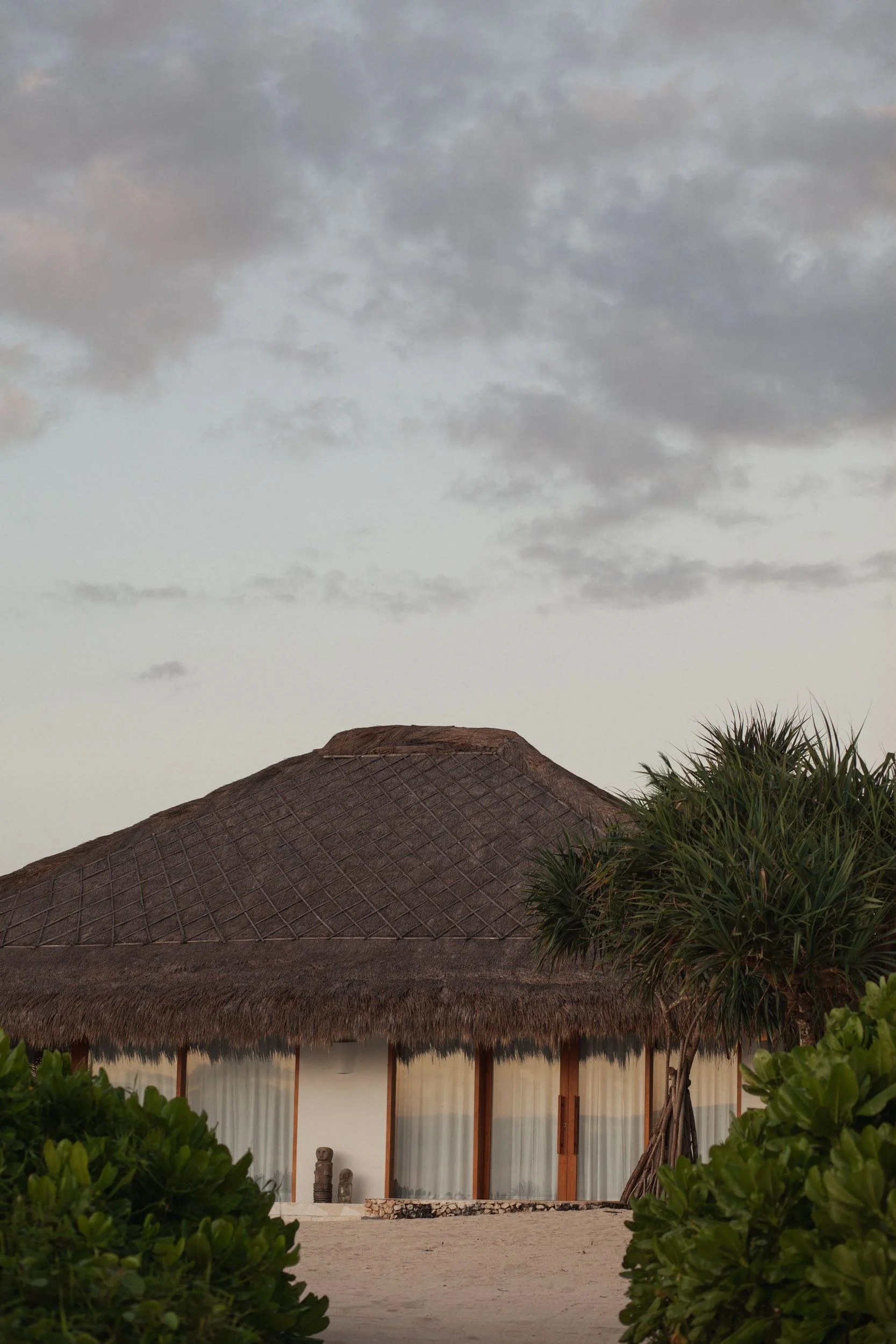 Thatched-roof building with outdoor patios, surrounded by green bushes, under a cloudy sky.