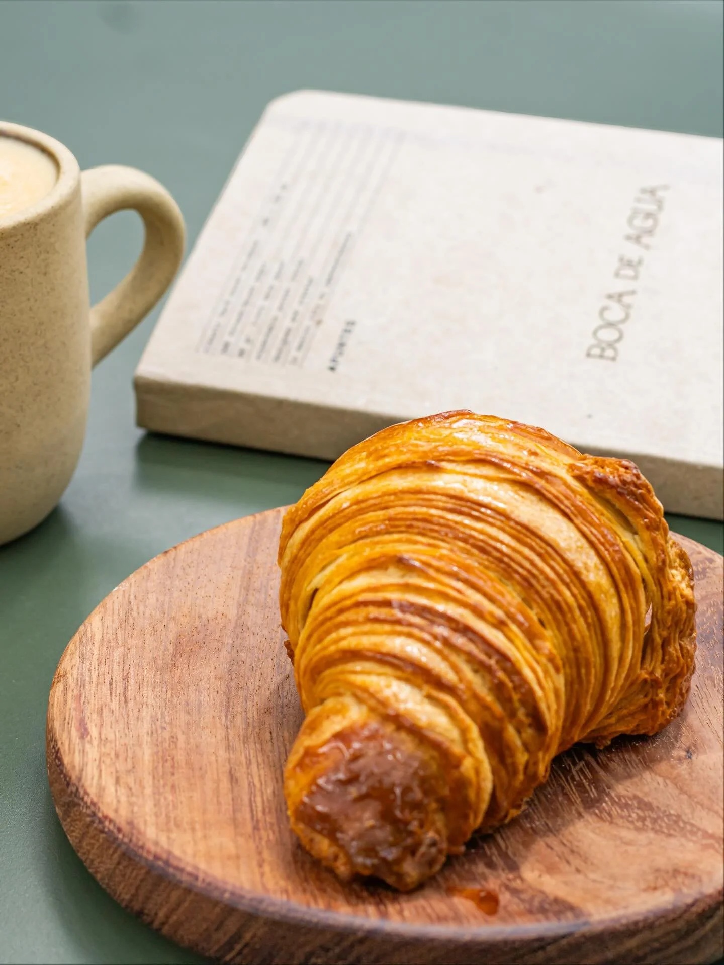 A croissant with a flaky, golden-brown exterior on a wooden plate, with a coffee mug and a closed book titled 'Book de Argia' in the background.