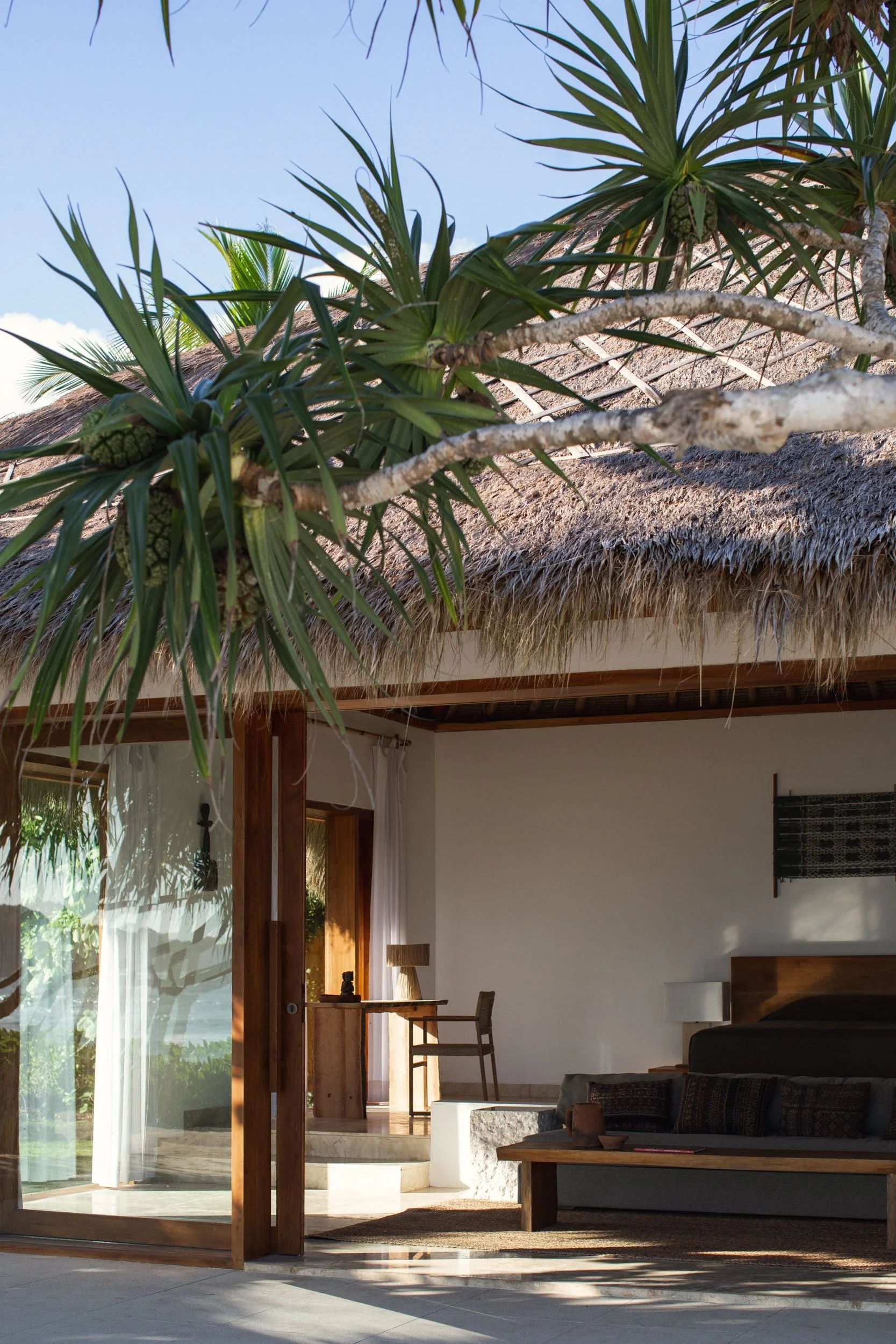 Interior of a modern home with a thatched roof, large glass doors, and a view of lush greenery outside. Modern furniture includes a gray sofa, wooden chairs, and a coffee table, with natural light flooding the space.
