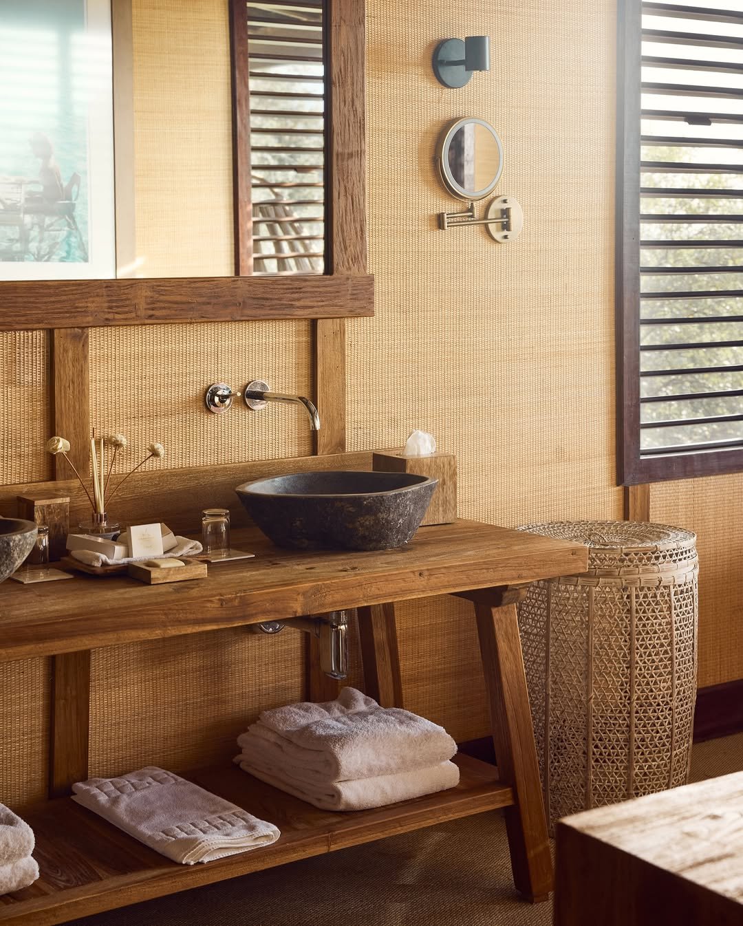 A rustic bathroom with a wooden vanity holding a black vessel sink, a mirror, and toiletries. Towels are neatly folded on a shelf underneath, with natural light coming through wooden window shutters.