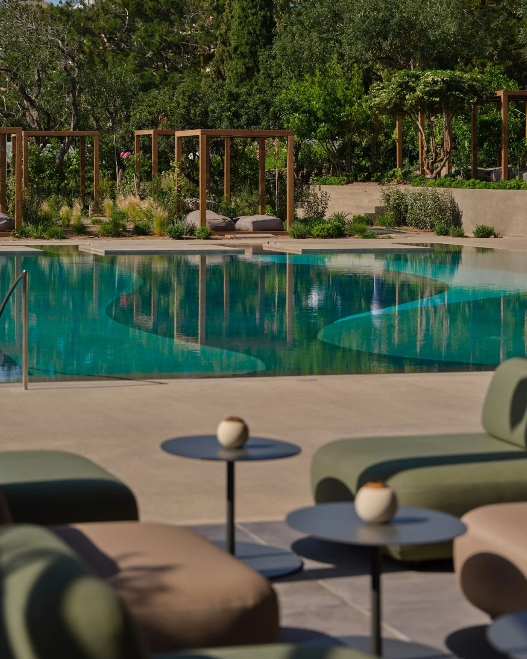 Outdoor pool area with lounge chairs and small tables, lush greenery, and wooden cabanas in the background.