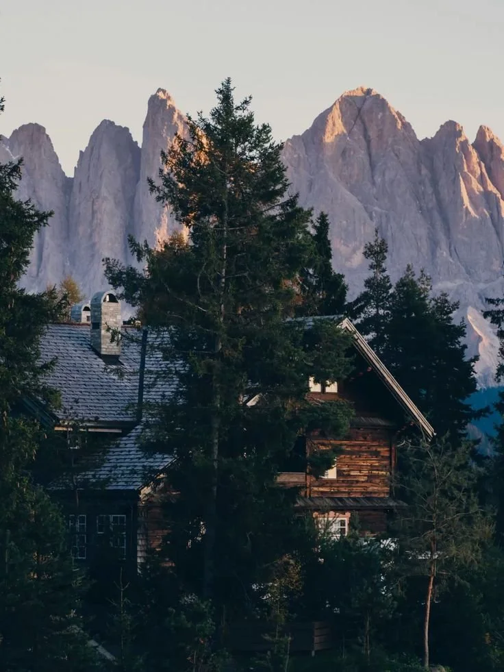 A mountain cabin surrounded by trees with tall rocky mountain peaks in the background during sunset.