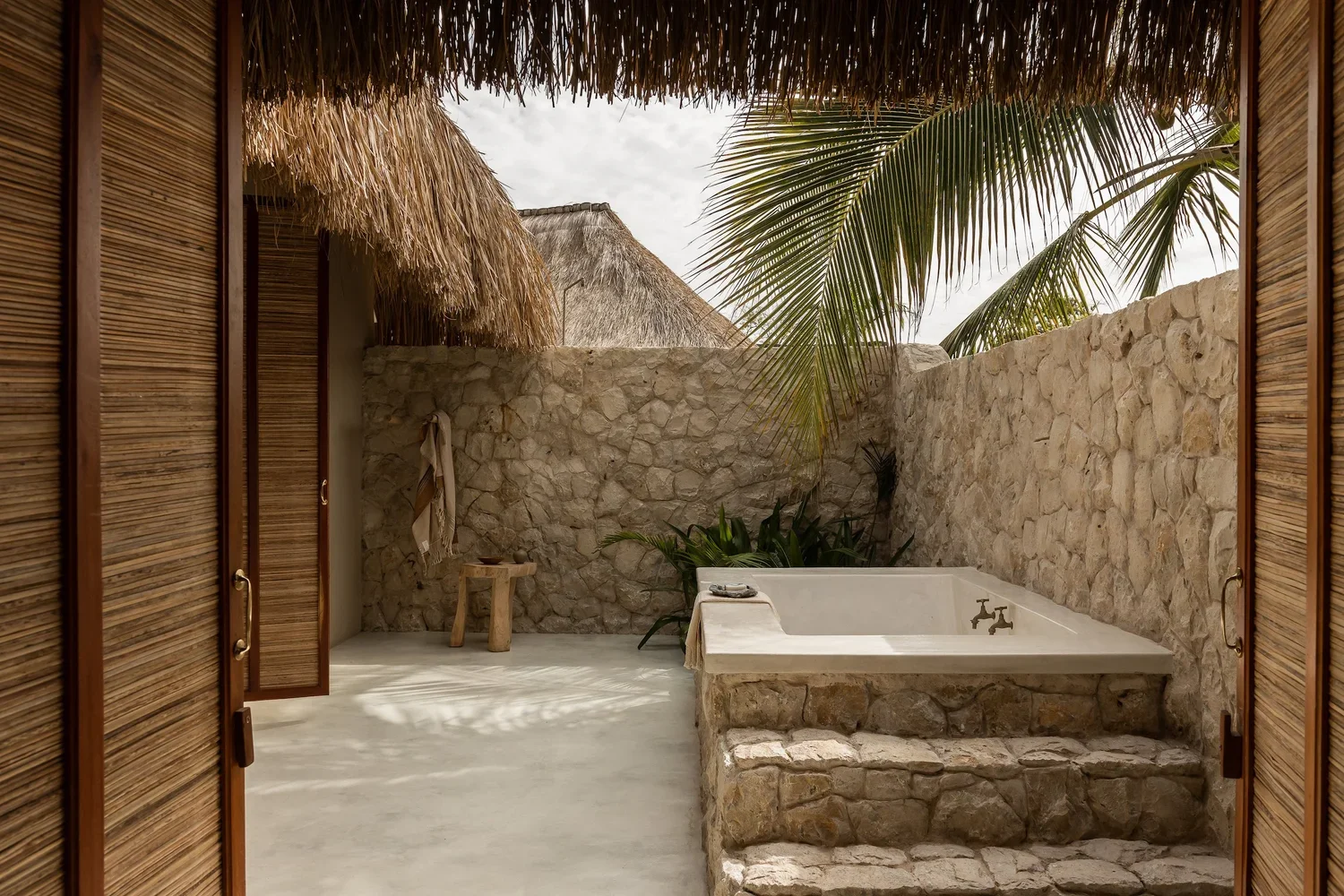 Open doorway leading to an outdoor stone bathtub with palm trees and stone walls in a tropical setting.