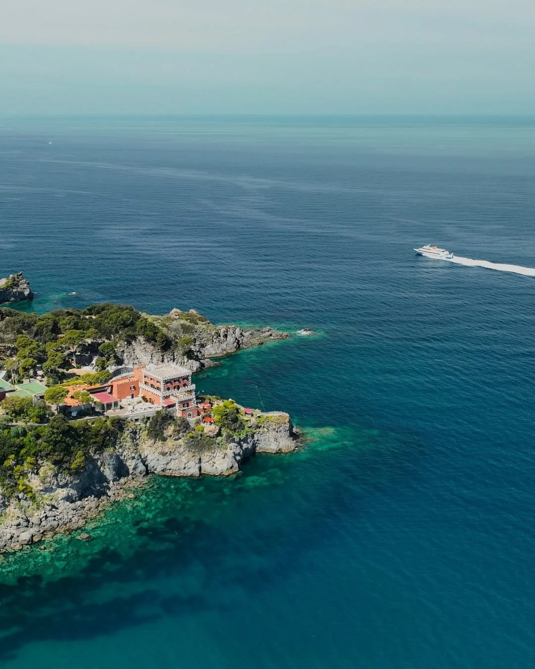 Aerial view of a coastal area with houses and greenery on rocky cliffs, and a large body of water with a boat speeding across it.
