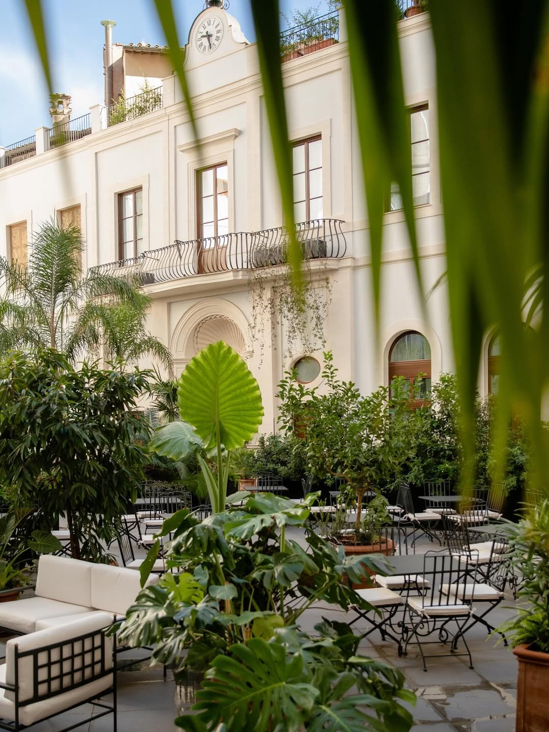 A lush garden courtyard with white outdoor furniture, potted plants, and greenery, enclosed by a white building with large windows and a clock on top, partly obscured by large tropical leaves in the foreground.