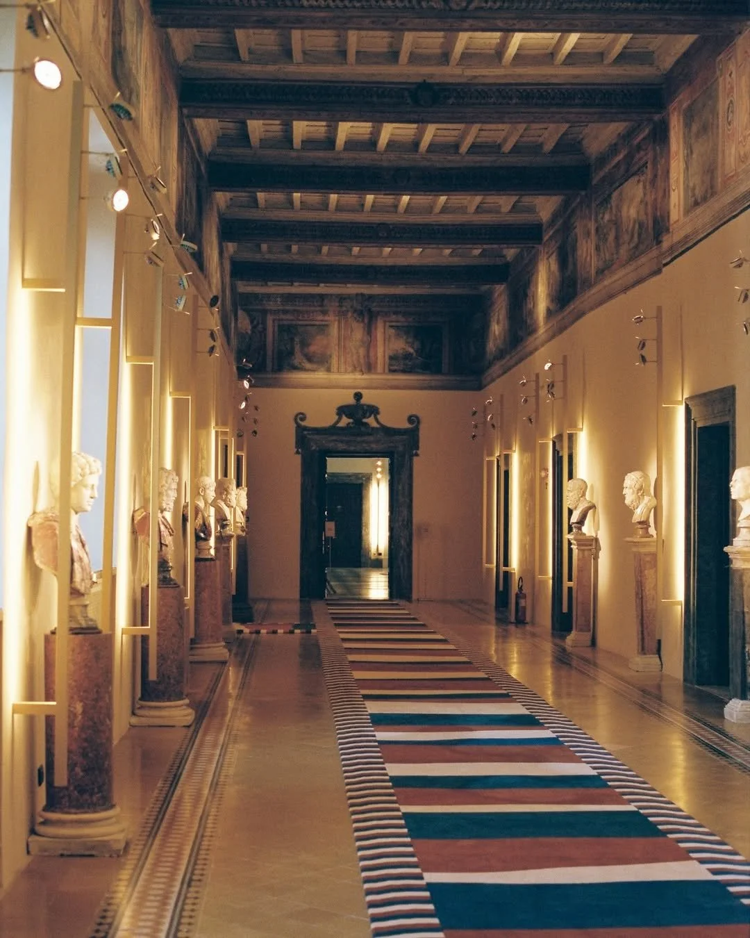 A long hallway in a museum or gallery with statues on pedestals lining each side, illuminated by wall-mounted lights, with a colorful striped carpet on the floor leading to a doorway at the end, and a decorative wooden ceiling.