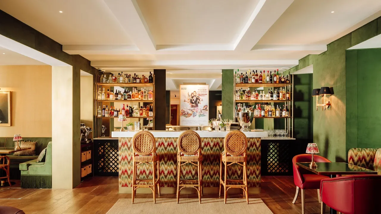 A stylish hotel bar with a patterned front bar counter, three wooden barstools, and shelves of liquor bottles in the background. There are green and red upholstered chairs and small tables with lamps, and warm ambient lighting.