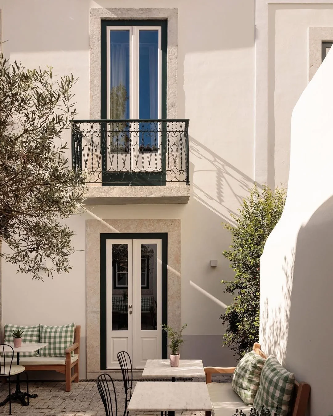 Outdoor patio area with white walls, a black metal balcony above, a set of white double doors with black trim, a wooden bench with green checkered cushions, a small white table with a pink potted plant, additional black chairs, and trees casting shadows on the walls.