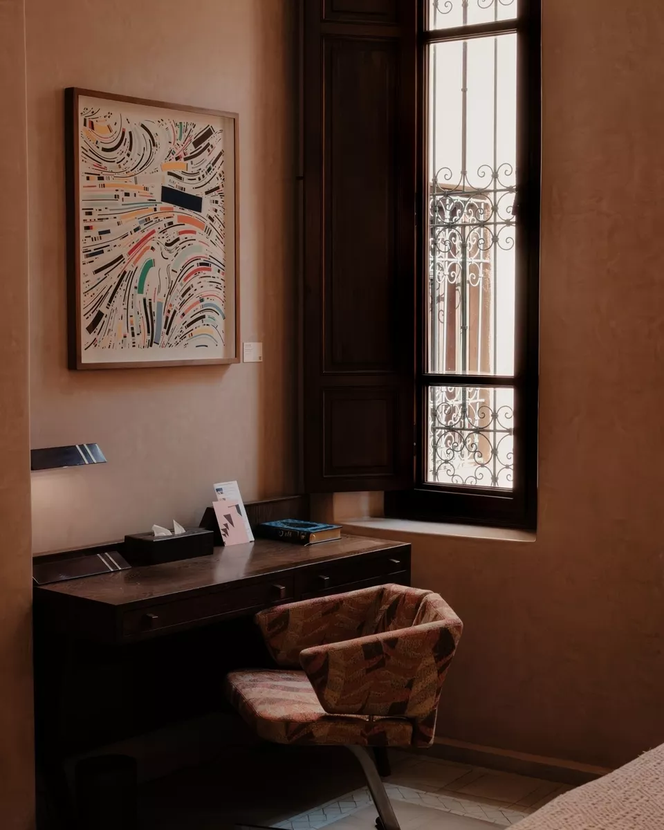 A cozy corner in a room with a window featuring decorative ironwork, a framed abstract artwork on the wall, a wooden desk with papers and a book, a tissue box, and a patterned upholstered chair.