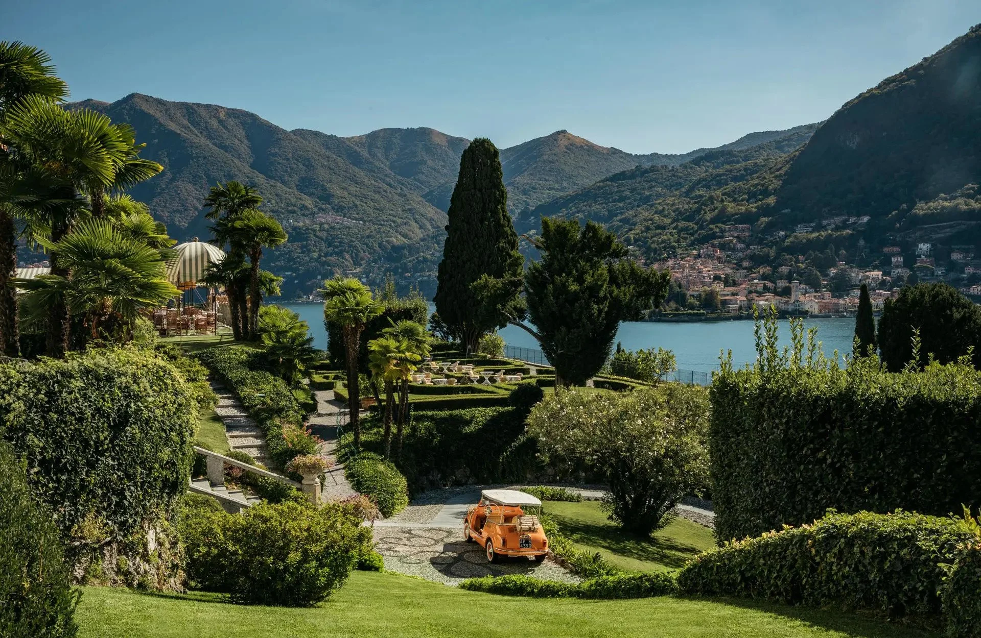 Lush green garden with steps leading down, a vintage orange car, tall trees, manicured hedges, a patio with striped umbrellas, and a lake with mountains in the background.