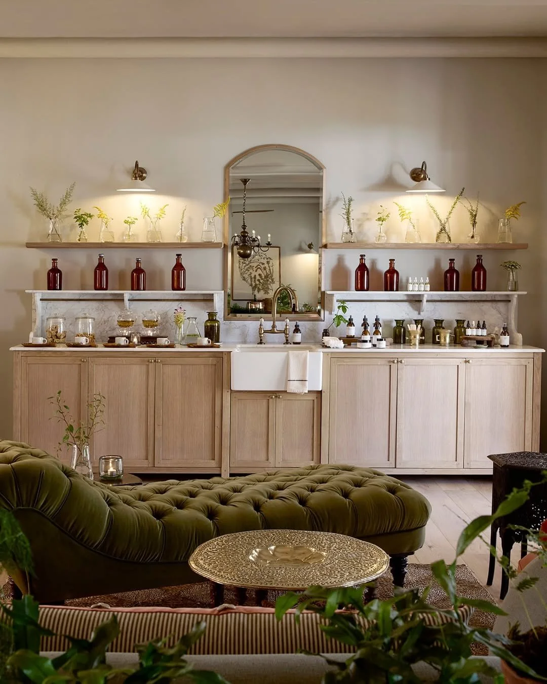 A cozy interior with beige cabinetry, a white farmhouse sink, and open shelves holding decorative bottles, glass jars with herbs and flowers, and various skincare or essential oil bottles. The space is accented with green plants, a tufted green velvet sofa, and a round ornate side table.