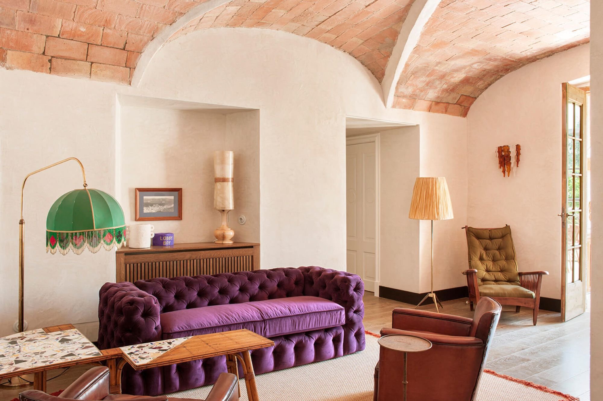 A cozy living room with a vaulted ceiling made of exposed brick and white walls. Features include a green vintage table lamp, a framed picture, a tall beige lamp, and a purple velvet tufted sofa. There's an armchair and a side table near a window that opens to outside greenery.