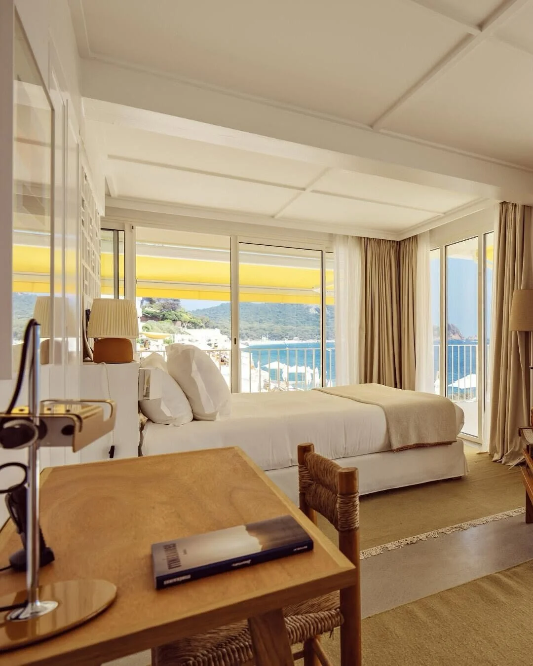 A bright hotel room with a large bed, white bedding, and multiple pillows, overlooking a balcony with a scenic view of the ocean and hills in the distance. There are beige curtains, a wooden desk with a lamp and a book, and wicker chair.