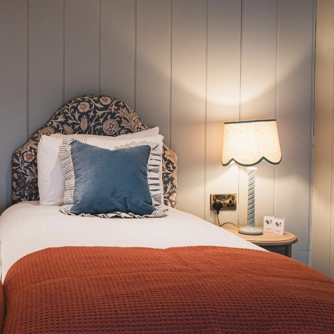A neatly made bed with a floral patterned headboard, layered with white pillows, a blue pillow with ruffled edges, and an orange blanket at the foot. A lamp with a twisted base and scalloped lampshade on a nightstand provides warm lighting.