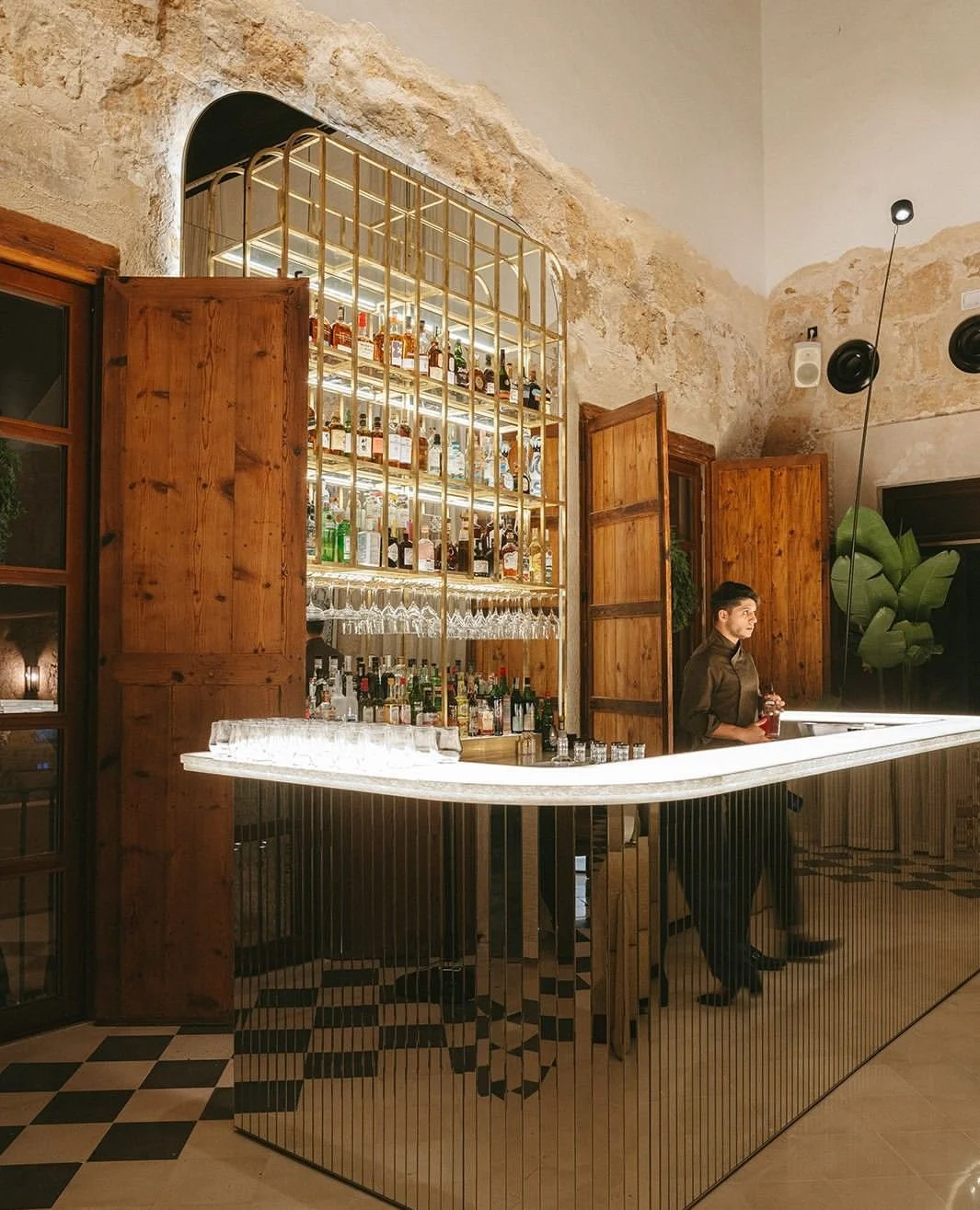 A bar with a curved, illuminated white countertop and a mirrored front, behind which bottles of liquor and glasses are displayed on shelves. An employee stands behind the bar, holding a drink, in a rustic interior with exposed stone walls and wooden shutters.