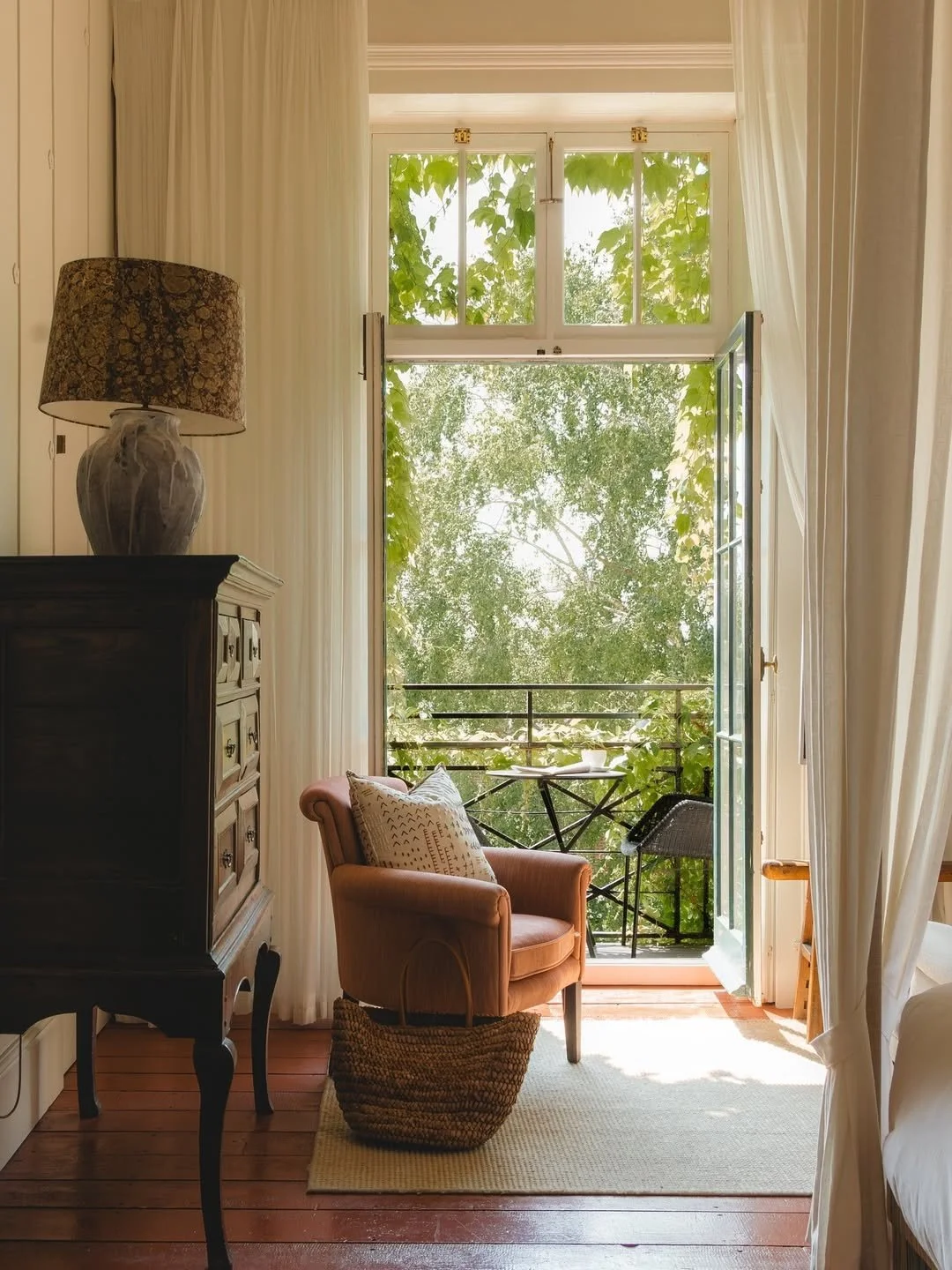 Sunlit room with open balcony door, pink armchair with patterned pillow, woven basket, vintage dresser with lamp, and view of trees outside.