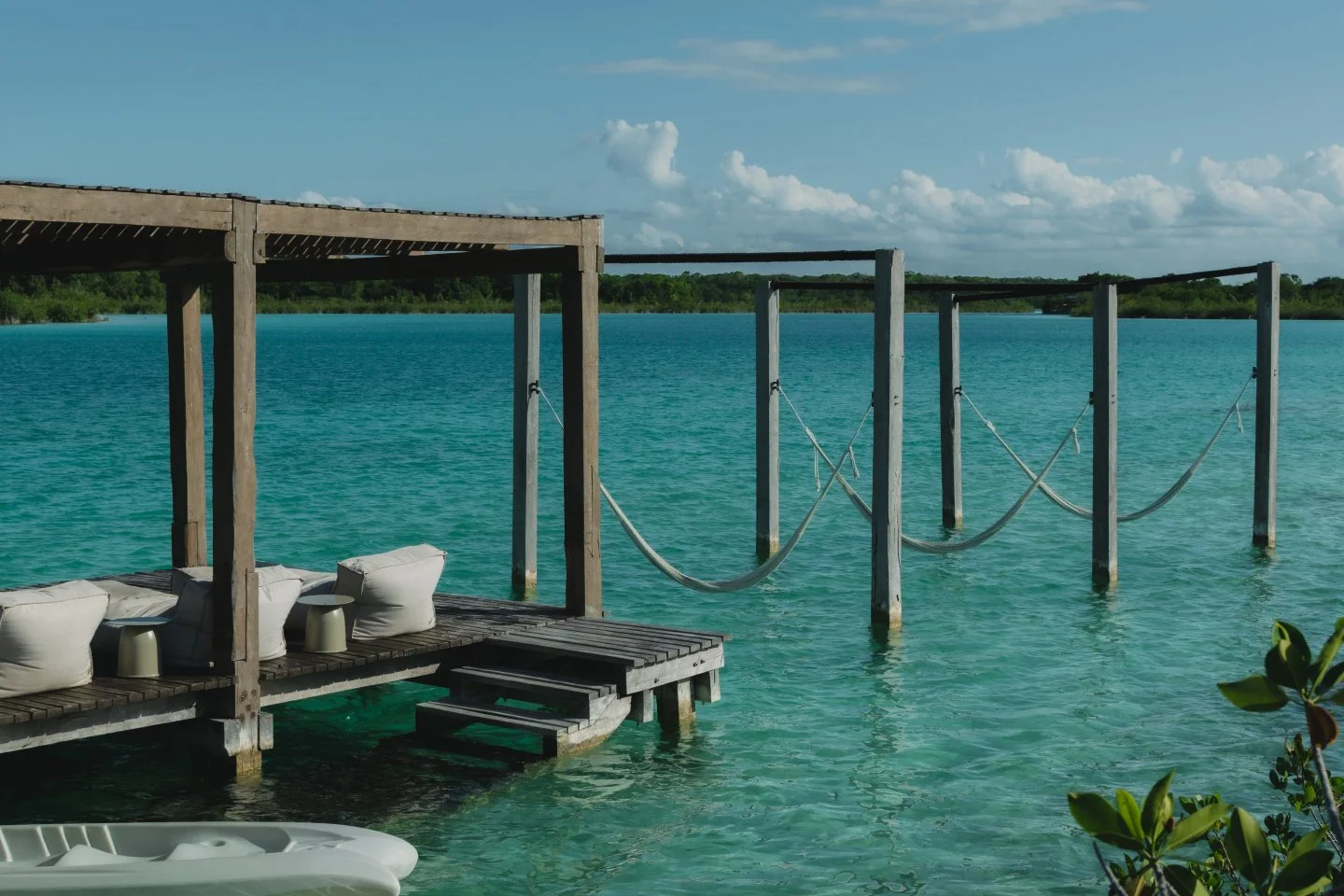 A wooden dock with a shaded seating area with white cushions and tables over a turquoise water body, connected to a second dock with rope barriers, with a distant green shoreline and partly cloudy sky.
