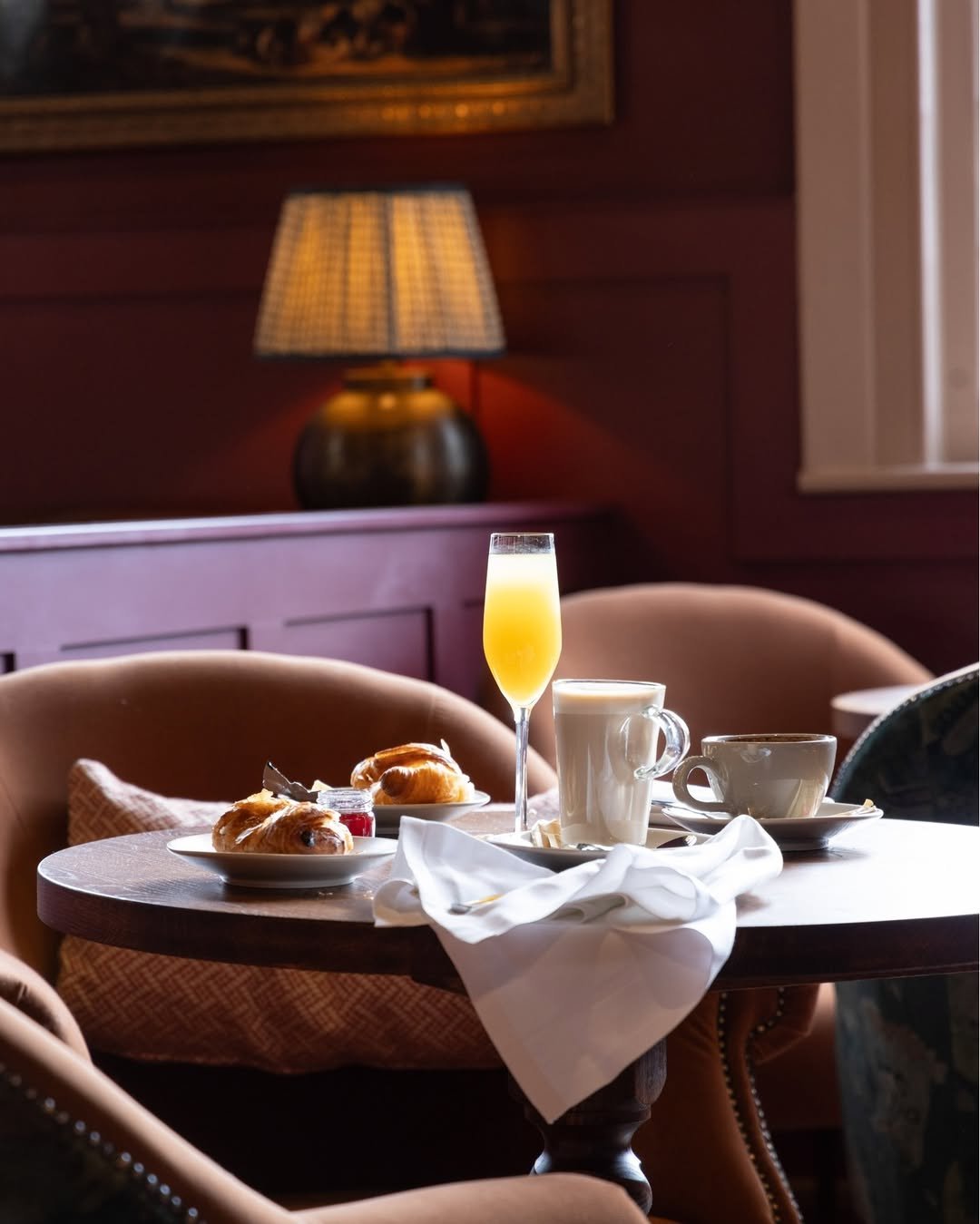 A table set for breakfast with croissants, a glass of orange juice, a mug of coffee, and a teacup in a cozy, dimly lit room with upholstered chairs and a lamp in the background.