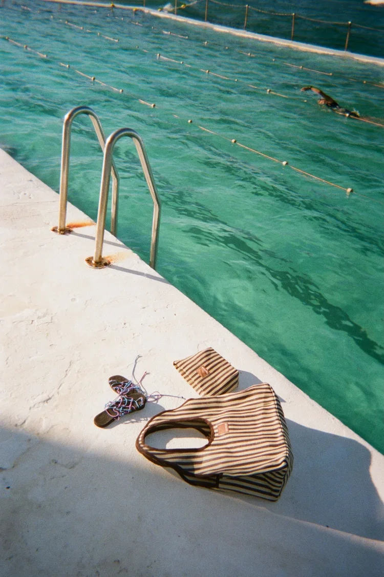 Stripes beach bag, a small matching pouch, and flip-flops on a poolside deck near a swimming pool with pool lane ropes and a person swimming.