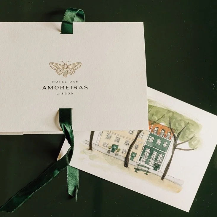 A white card with a green ribbon, featuring a butterfly logo and text: 'Hotel Das Amoreiras Lisbon', resting on top of a watercolor illustration of a street with trees and buildings.
