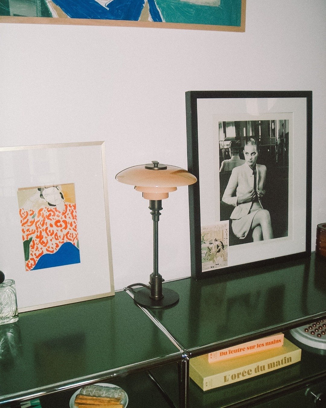 A green metallic surface with framed artwork, a vintage lamp, a small glass jar, and books on it. One of the framed artworks features a black-and-white photograph of a woman sitting, holding a cigarette. The books are titled in French.