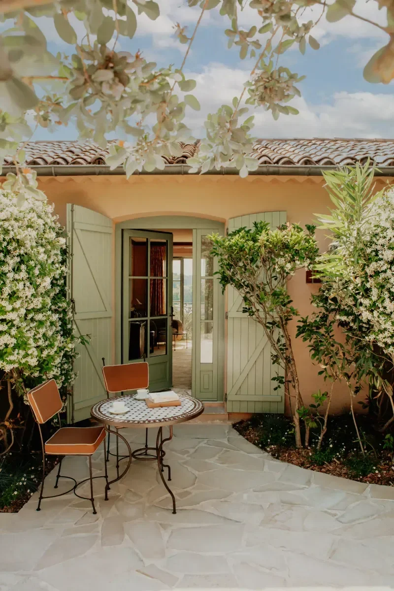 A cozy outdoor patio with a small round table, two chairs, and open green shutters leading into a house, surrounded by lush green bushes and trees, under a partly cloudy sky.