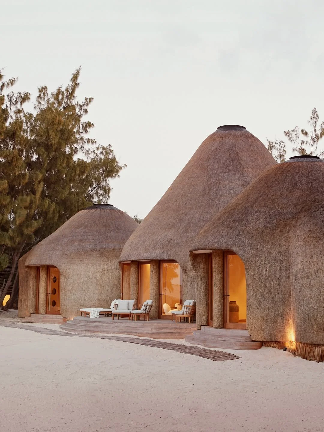Three grass-thatched round huts with large windows and outdoor seating on a sandy surface, trees in the background, and a clear sky.
