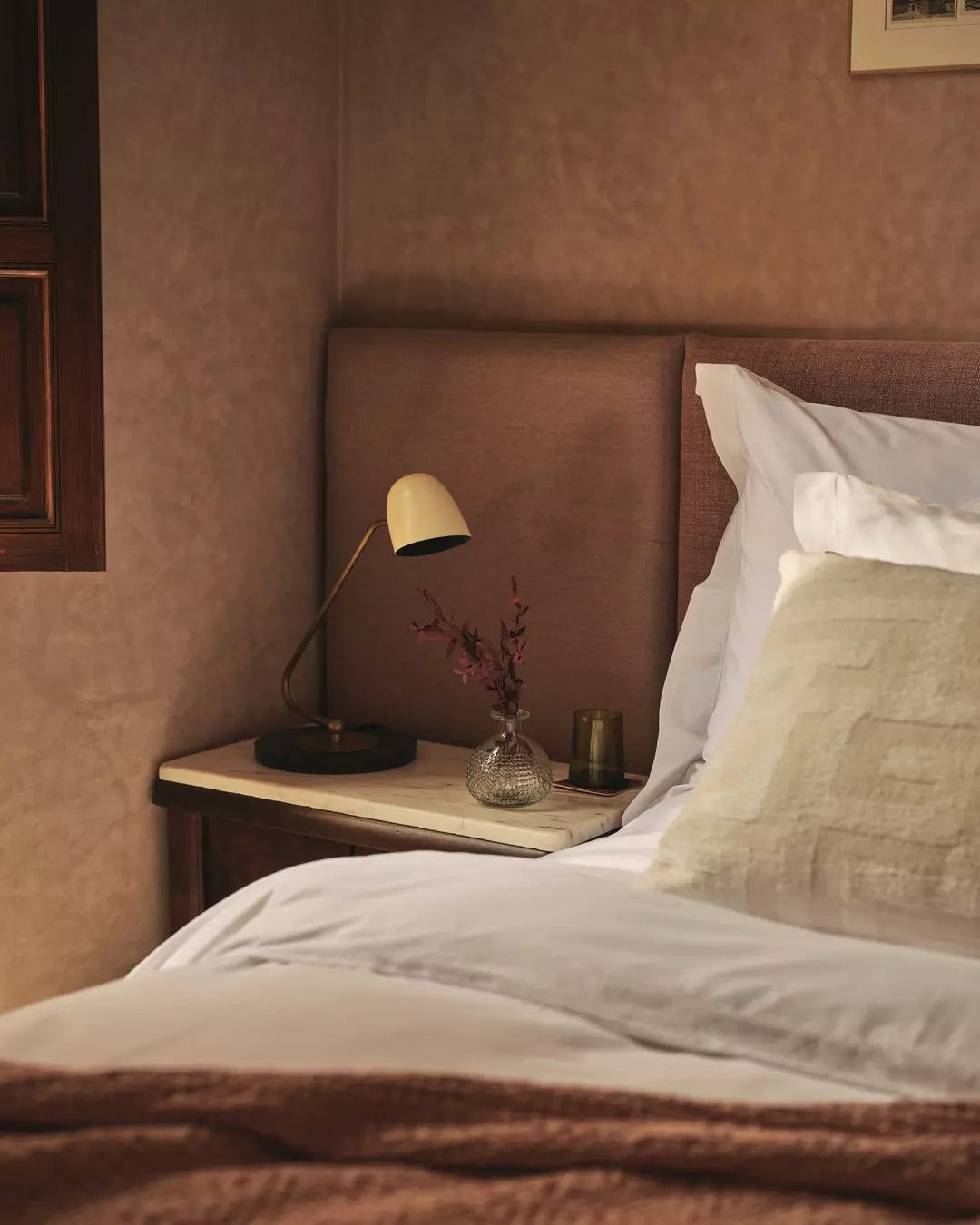 A cozy hotel bedroom with a bed, beige pillows, a beige upholstered headboard, a nightstand with a cream marble top, a modern yellow desk lamp, a small glass vase with pink flowers, a dark glass candle holder, and a textured wall.
