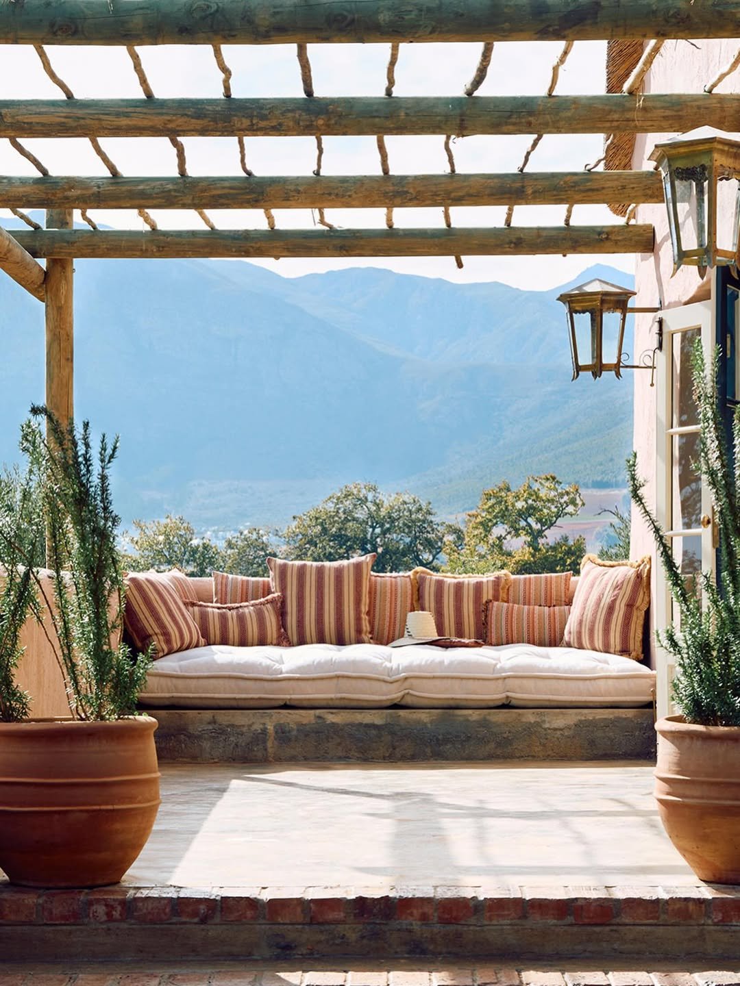 A cozy outdoor patio with a cushioned daybed filled with striped pillows, potted plants, and a panoramic view of mountains and trees in the background.
