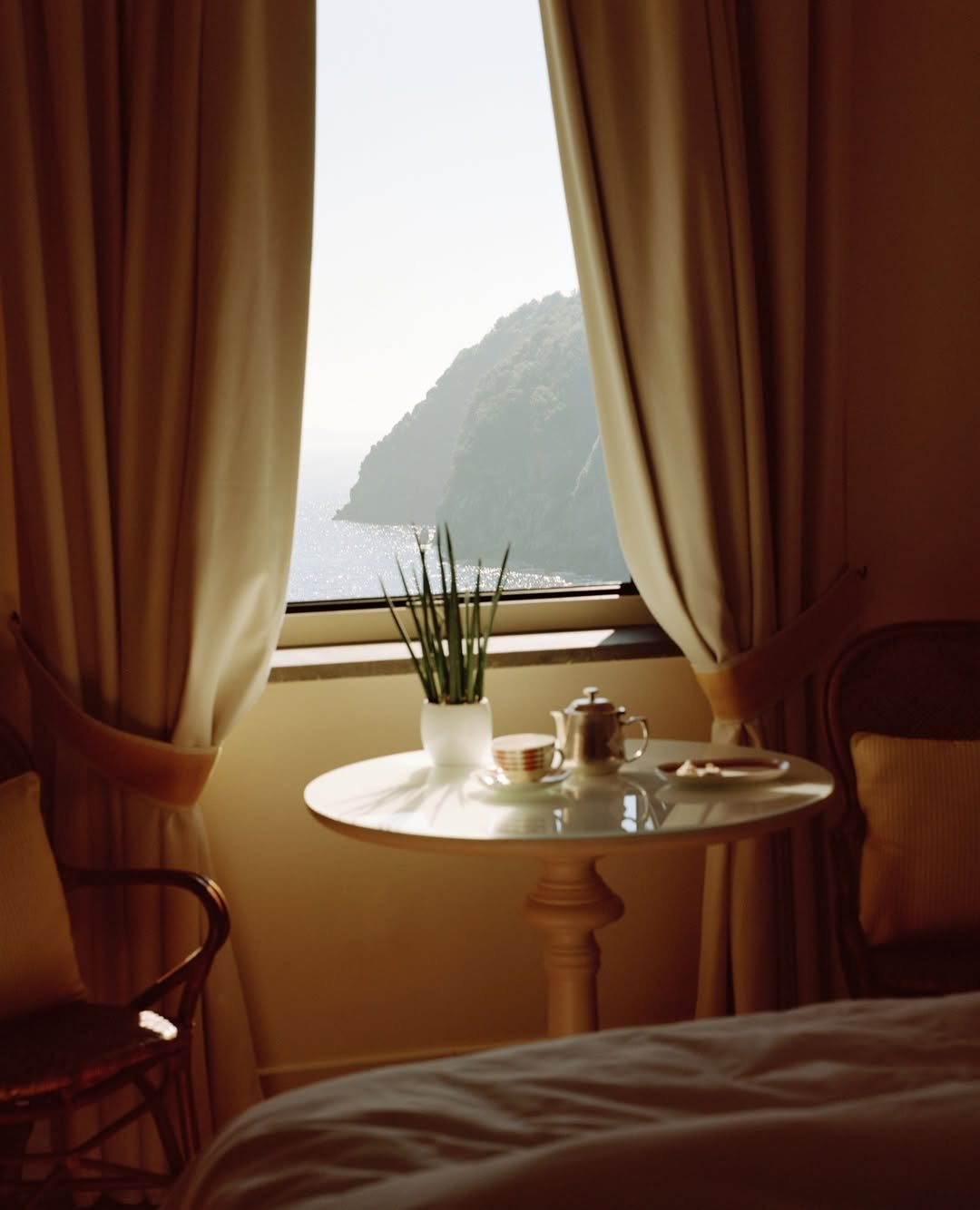 Sunlit window with beige curtains revealing a scenic view of a mountain and water, a small round table with a potted plant, teapot, teacup, and plate in a cozy, elegant room.