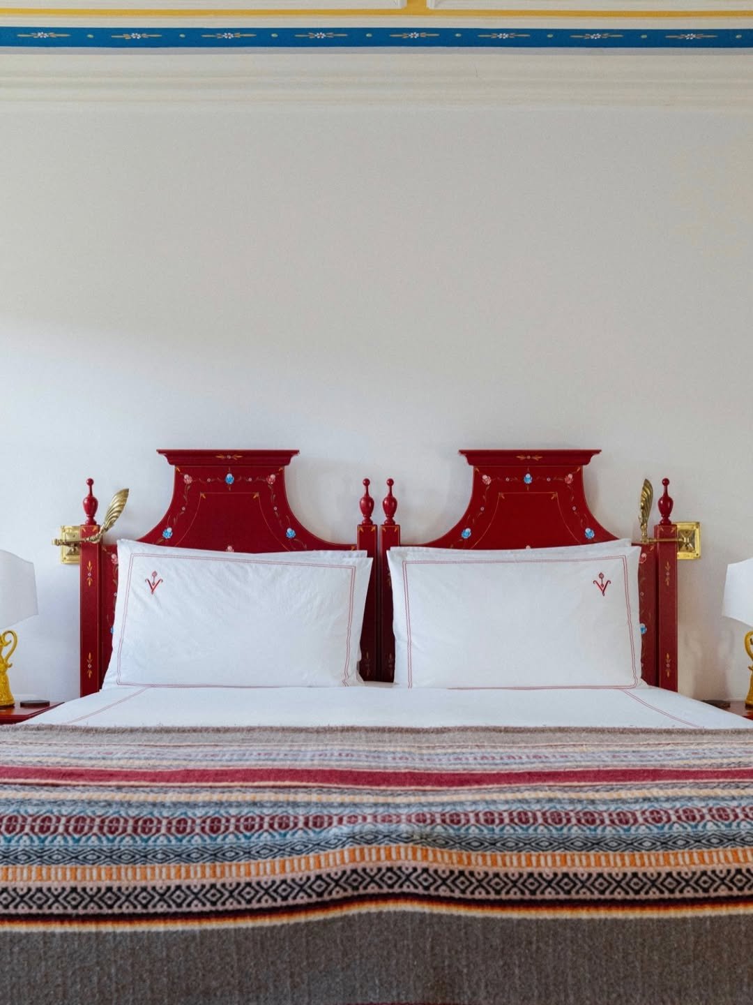 A bedroom with a bed featuring two white pillows with red embroidered initials, a red wooden headboard with decorative blue accents, and matching red bedside lamps, on a bed with a multicolored patterned blanket.
