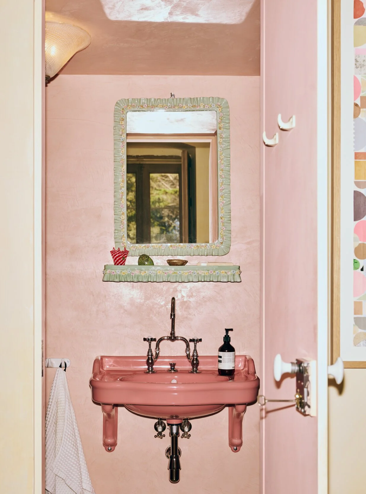 A small pink vintage sink with a mirror above it on a pink wall. There are decorative items on the mirror shelf, and a soap dispenser on the sink. A towel hangs on a hook to the left.