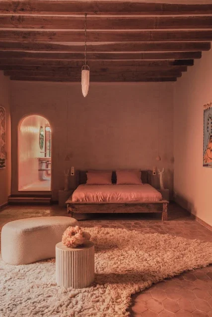A bedroom with a wooden ceiling, a bed with pink bedding, a fluffy rug, and a curved bench with a flower beside it.