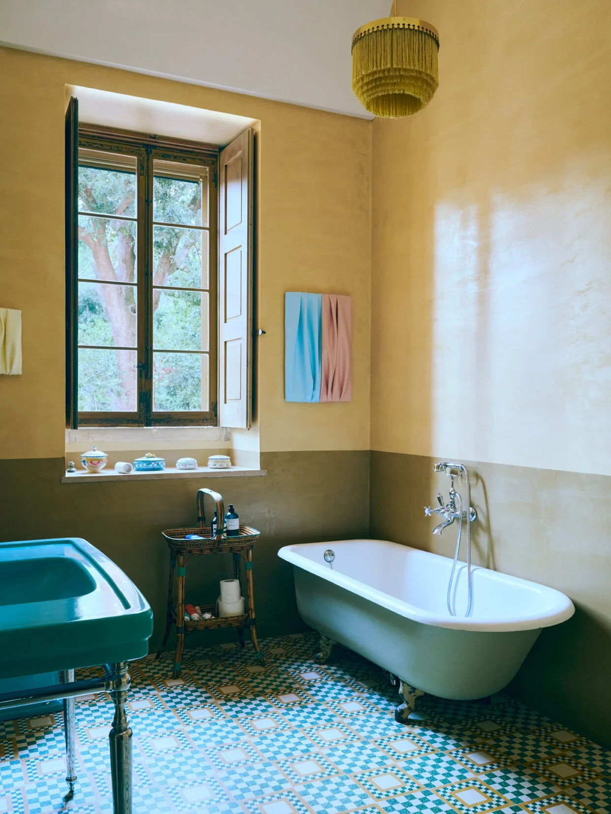 A vintage-style bathroom featuring a white clawfoot bathtub, a green sink, a wicker side table with toiletries, an open window with outdoor trees, a yellow hanging light, and a pastel towel hanging on the wall.