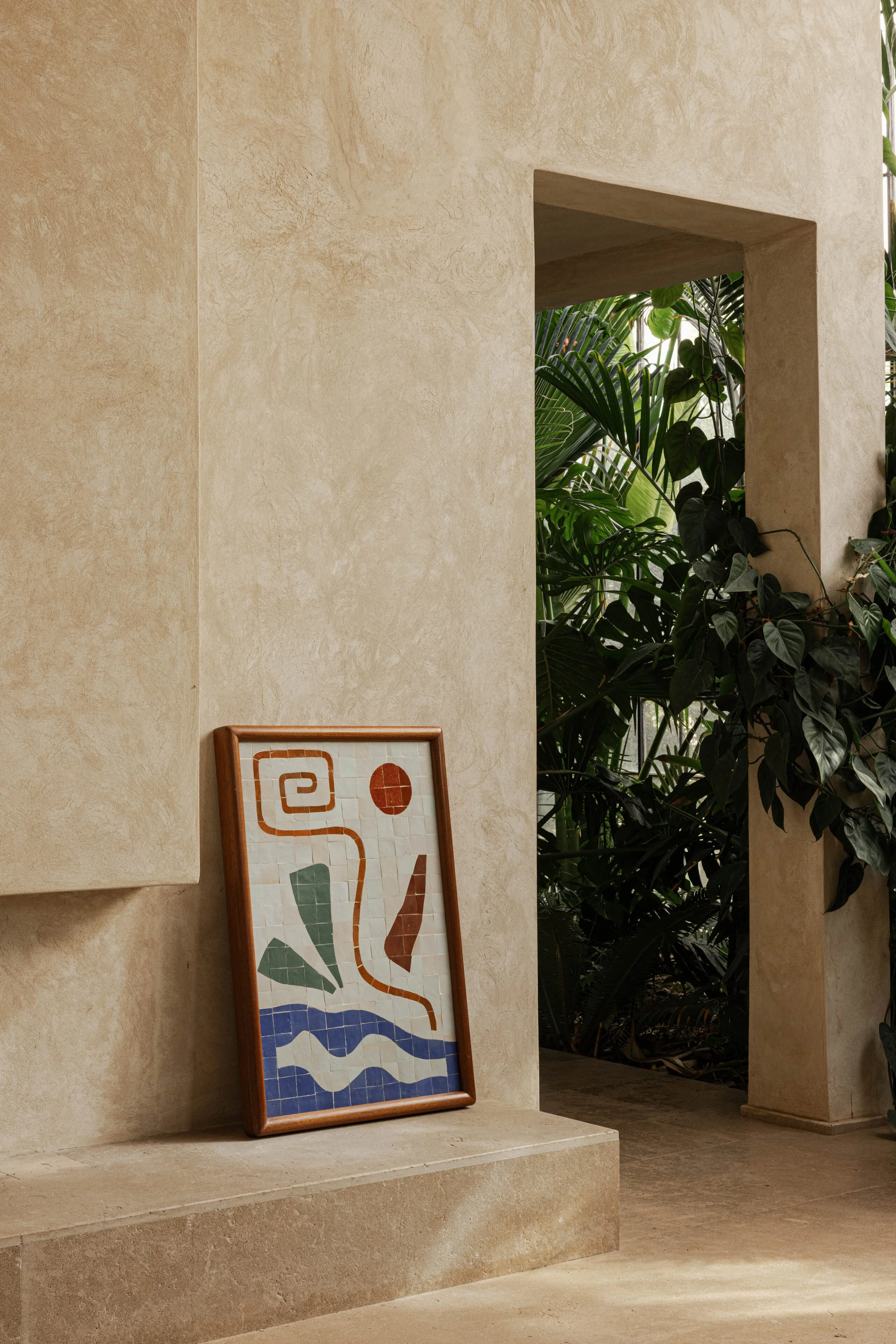 Interior view of a beige stone wall with a framed geometric artwork resting on a ledge, and lush green plants visible through a square opening.