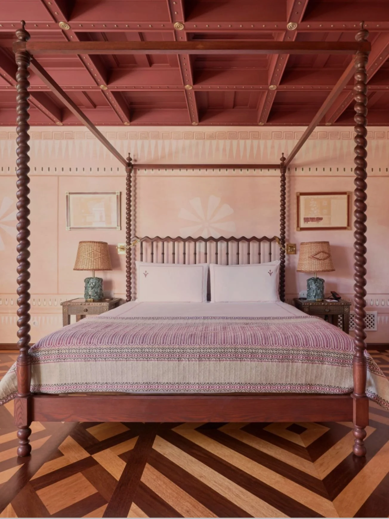 A luxurious hotel room with a four-poster bed featuring carved wooden posts, placed on a patterned wooden floor. The bed has white bedding and two matching pillows. On either side of the bed are wicker nightstands with table lamps and decorative bases. The wallpaper is pink with geometric patterns, and there are framed pictures on the wall. The ceiling has a wooden grid design.