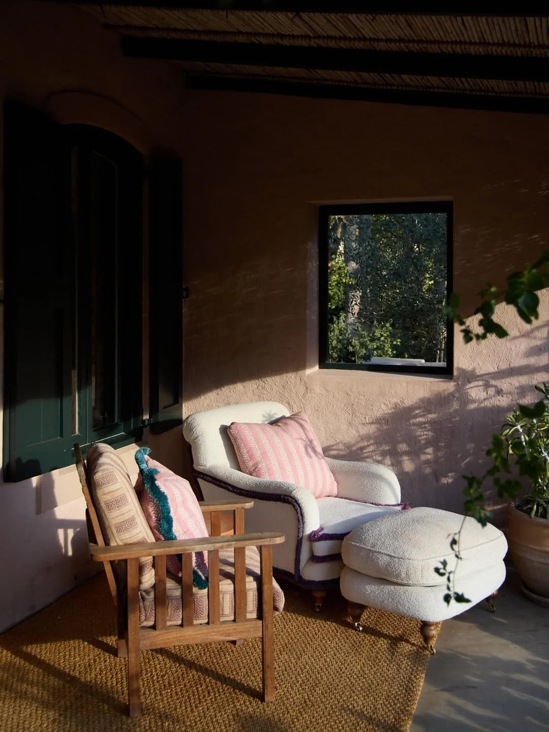Sunlit porch area with two vintage armchairs, one wooden with patterned cushions and the other upholstered in white with pink striped pillows, paired with a matching ottoman, on a textured rug next to a potted plant, with a window showing trees outside.