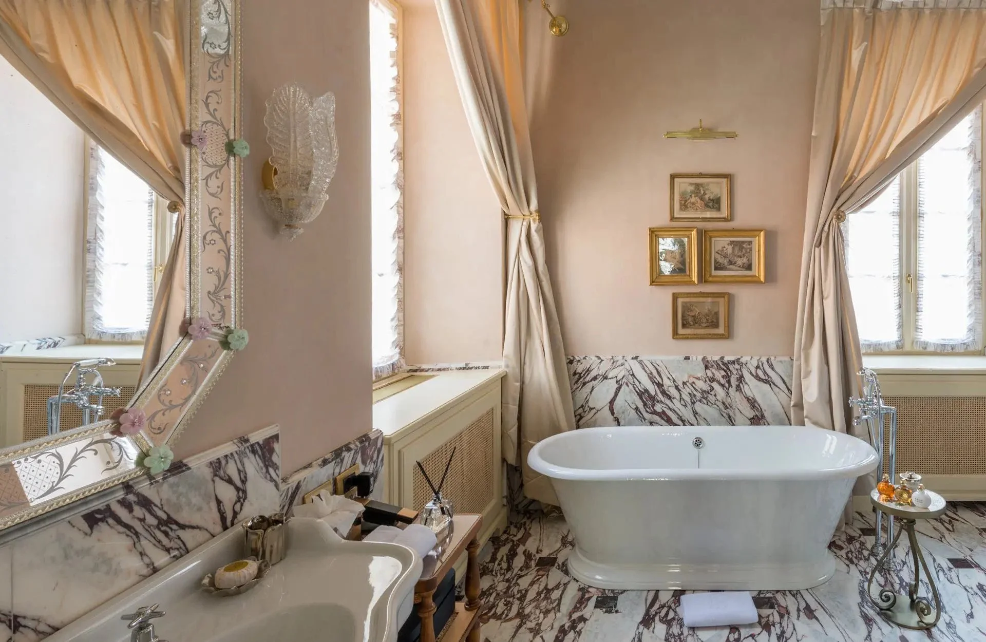 A luxurious bathroom featuring a large freestanding bathtub with marble flooring and walls, surrounded by elegant curtains, framed artwork, and vintage-style fixtures, creating a classy and sophisticated ambiance.