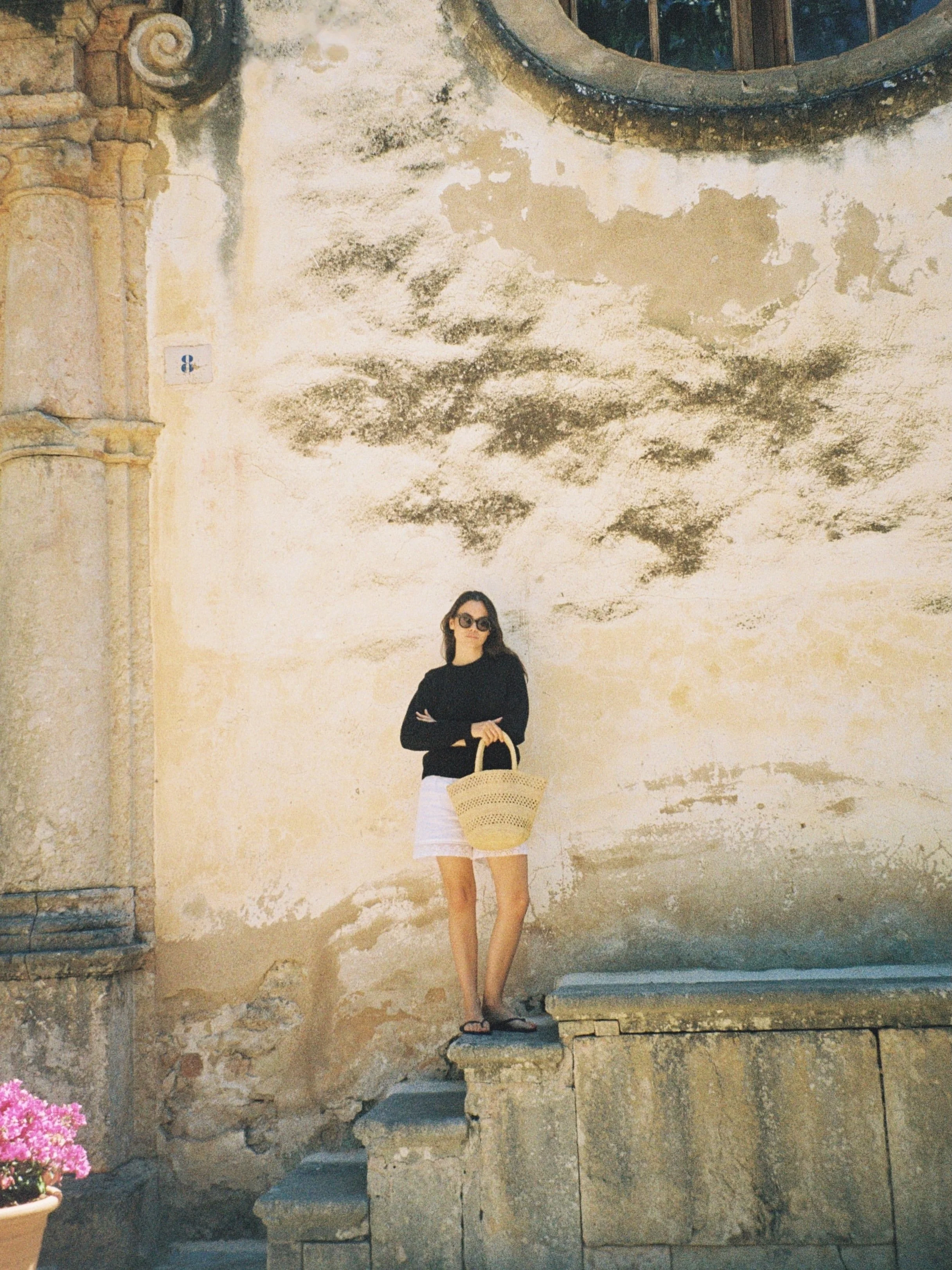 A woman with long dark hair wearing sunglasses, a black top, a white skirt, and sandals, standing on a stone step in front of an old, weathered wall with a large circular window. She is holding a woven straw bag. The wall has patches of black mold and peeling paint. To the left is a potted pink flower.