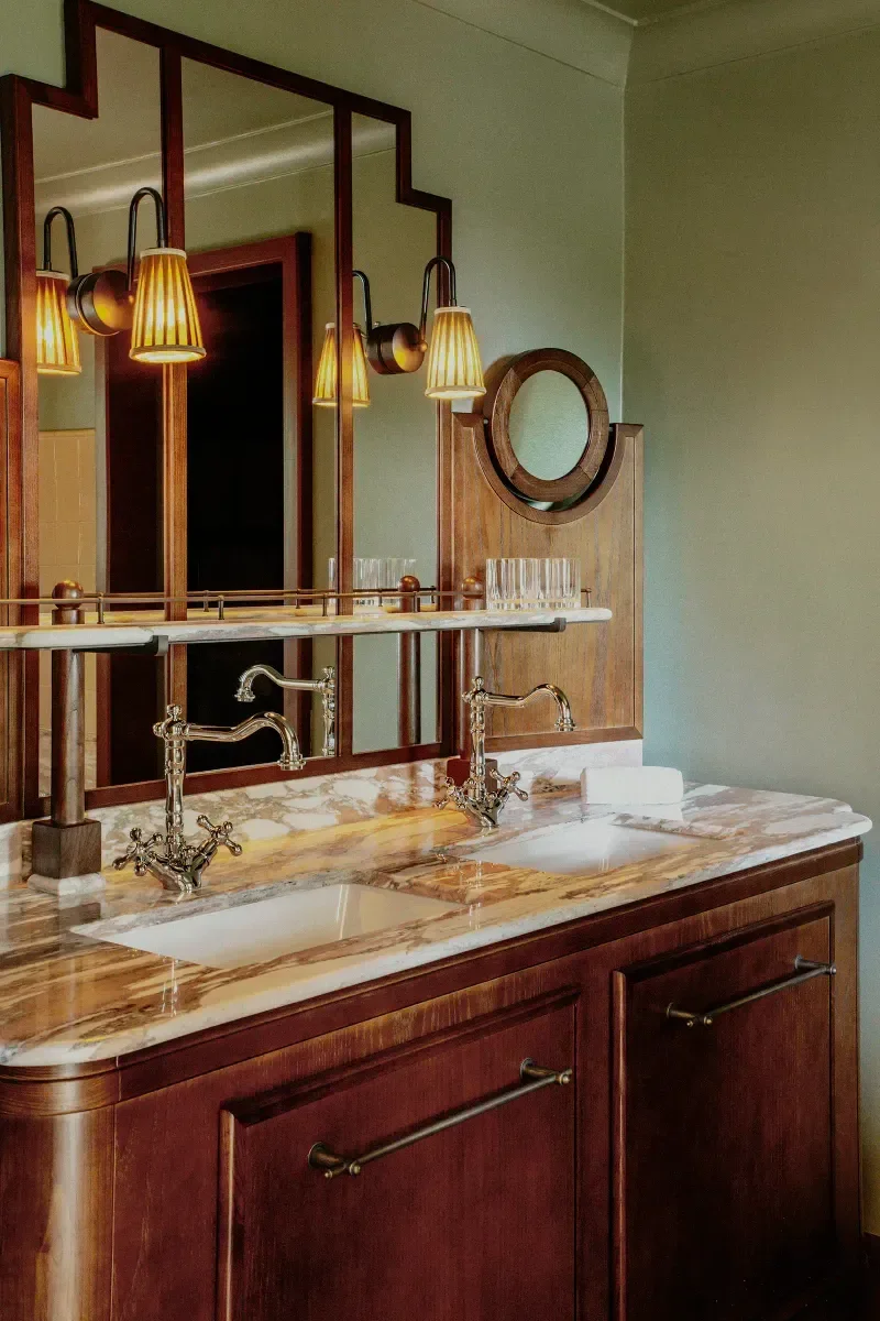 A vintage-style bathroom vanity with double sinks, brass faucets, and a marble countertop, reflected in a large mirror with brass sconces and a decorative wooden frame.